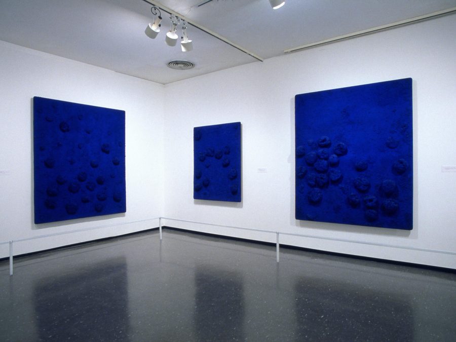 Installation of three blue paintings with objects attached to them in the corner of a gallery