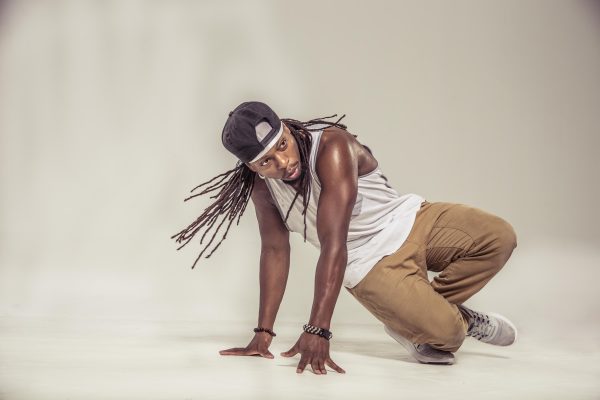 A male-presenting person with dark skin shown in the middle of a break dancing move, their dreads swaying from the movement, in an all white room