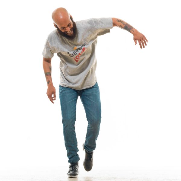 A male-presenting person with light brown skin, a shaved head, and a full brown beard, in the middle of a tap dance move against an all-white background