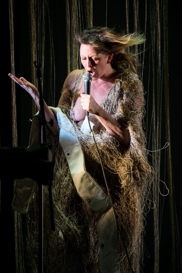 A woman bends her head toward a microphone she holds in her left hand, her mouth wide open. She wears a fringe covered robe which opens slightly to reveal her right breast