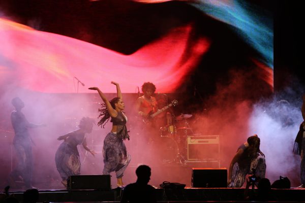 A female-presenting person dances across a smoky stage, their arms up in the air, with a band behind them. The entire scene is cast in a pink light.