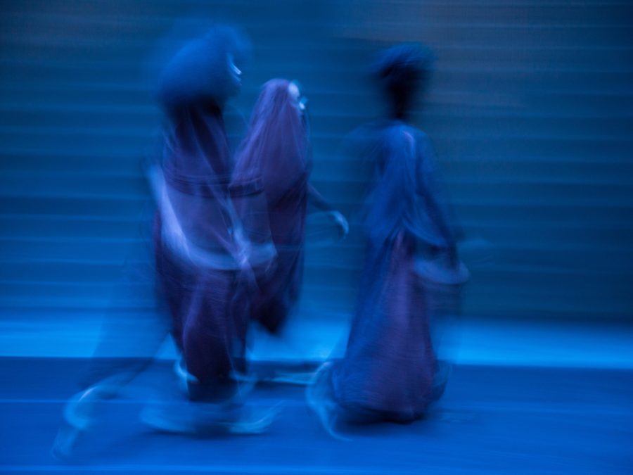 In this blue-toned photograph, three blurry, ghostlike figures in long dresses or robes walk toward the right side of the frame.