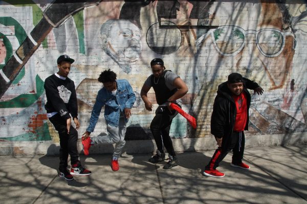 Four young men dance on a city sidewalk in front of a mural on a brick wall.