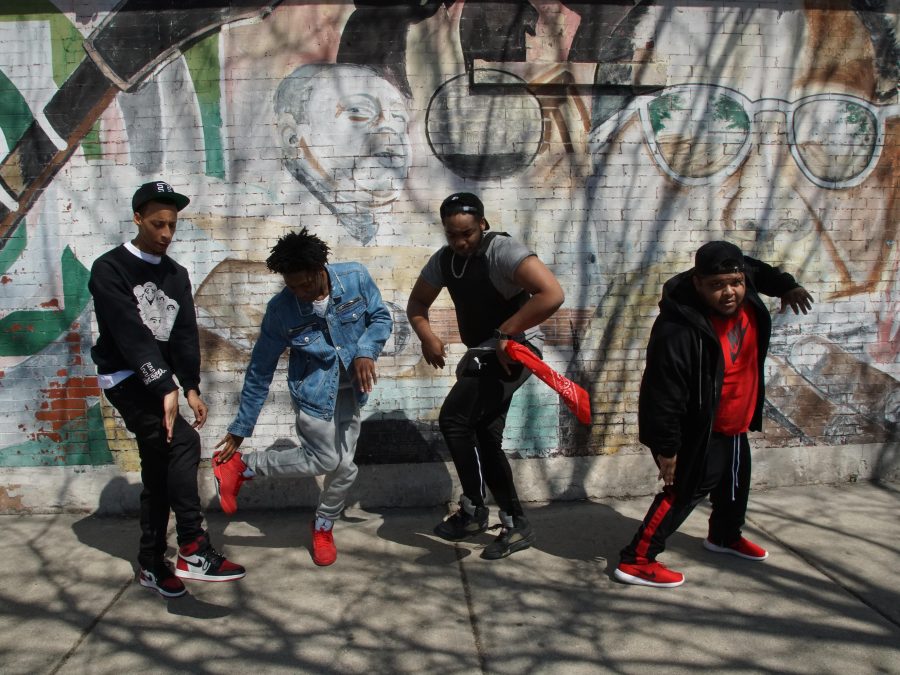 Four young men dance on a city sidewalk in front of a mural on a brick wall.