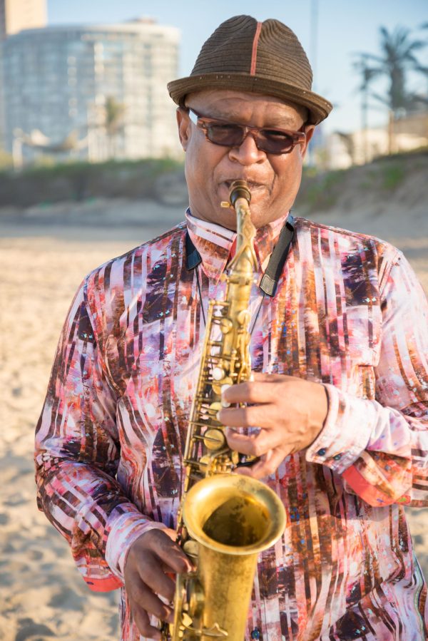 An older man holds a saxophone on a tropical beach on a sunny day.