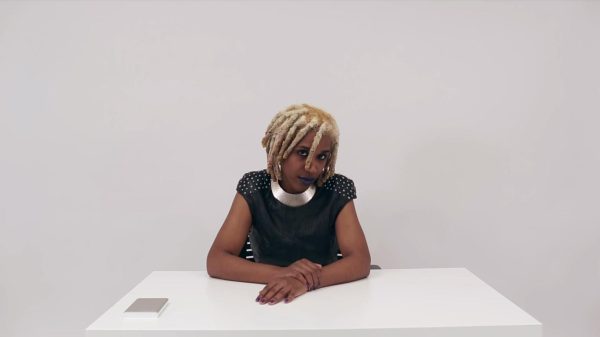 A female-presenting person with light brown skin and blond dreadlocks sits at a white table against a white backdrop