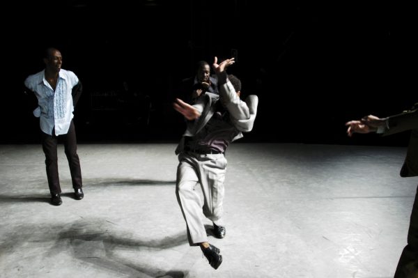 Three people on a dark stage. The person in the foreground jumps in the air.