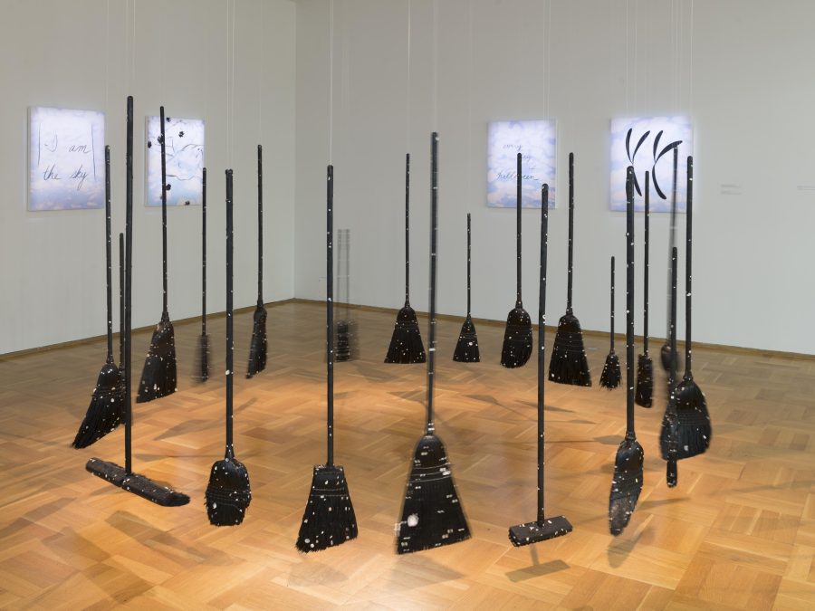 Various types of brooms hang in a circle in a gallery space