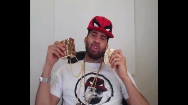 A male presenting person wearing a Spiderman hat and a shirt of himself in the hat holds up two large medallions attached to gold necklaces around his neck
