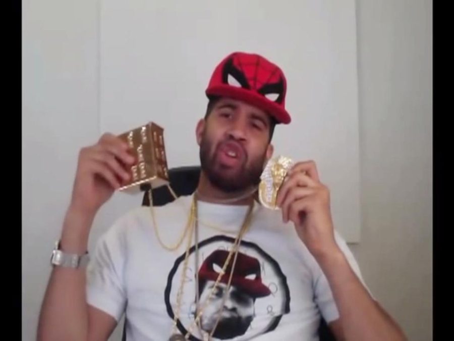 A male presenting person wearing a Spiderman hat and a shirt of himself in the hat holds up two large medallions attached to gold necklaces around his neck