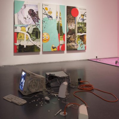 Pieces of an old desktop computer are strewn about the floor of a gallery with other artworks hanging on the walls nearby