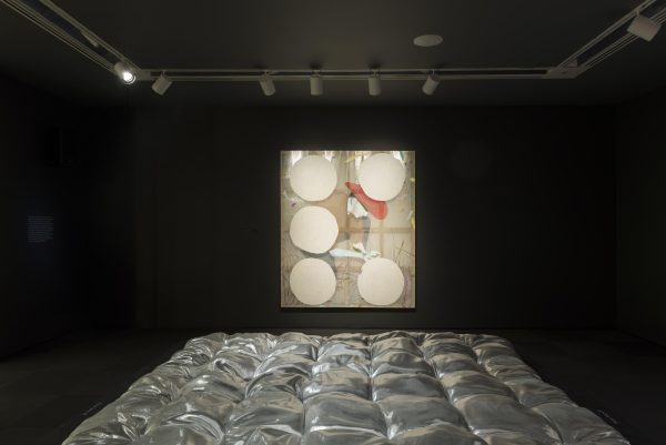 A darkened gallery features a metallic silver blanket in the foreground and a painting of a cowboy crouching in the background.