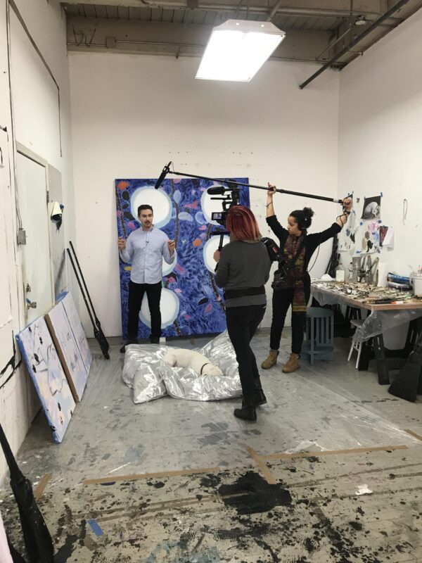 Two young feminine people hold recording equipment while a young dark-haired man speaks. They are in an artist's workspace with various artworks.
