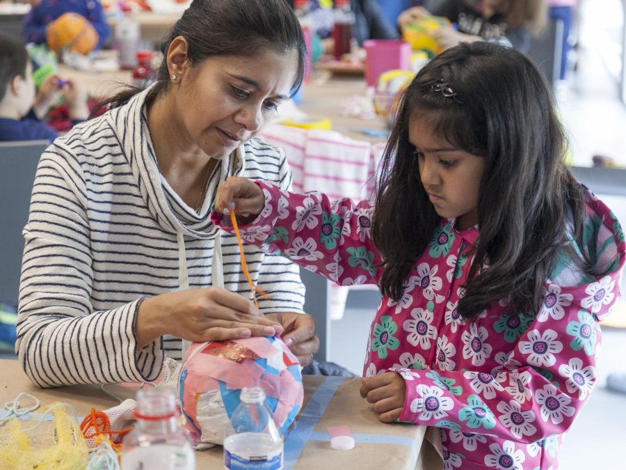 A child in a pink floral coat wraps an orange string around a colorful paper ball. An adult holds the string in place, assisting the child.