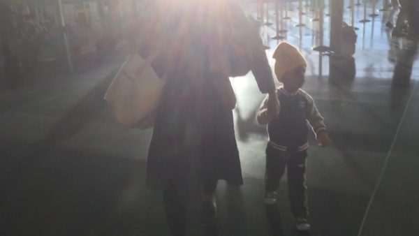 A sun glare obscures the backs of an adult leading a small child through a room filled with stanchions.