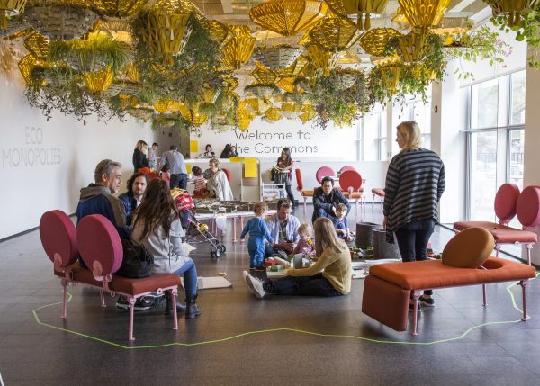 Adults and childrens sitting on the floor and pink chairs in a bright room filled with hanging plants