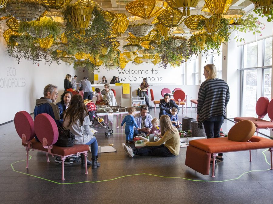 Adults and childrens sitting on the floor and pink chairs in a bright room filled with hanging plants