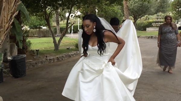 A dark-skinned woman with her hair and makeup done wears a sleeveless wedding dress with a long train, held by another person behind her. She holds up the dress and looks down toward the path ahead of her.