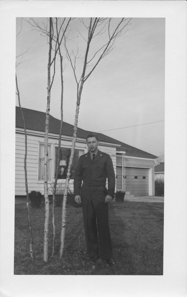 old black and white photograph of a man in an army uniform standing next to a birch tree outside a ranch-style house