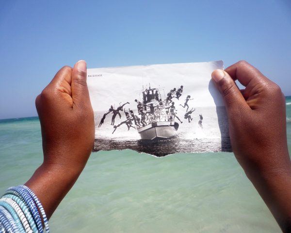 Two brown-skinned hands hold up a black and white ripped photo of people fleeing a boat over a body of water.