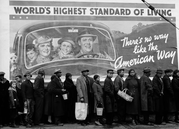 Black and white photo of a line of people under a painted billboard exclaiming World's Highest Standard of Living with a family of four picture and a tagline in cursive: There's no way like the American Way.