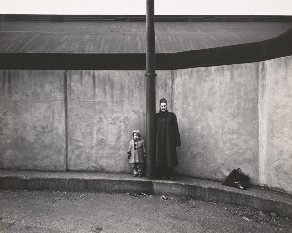 A woman and child in coats stand against a concrete structure