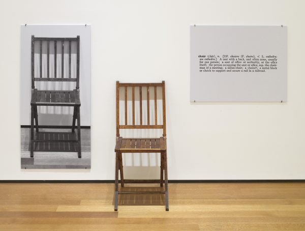 A black and white photograph of a chair, a wooden chair, and a printed panel of text featuring the dictionary definition of a chair appear in a row in a room with a wooden floor and a white wall