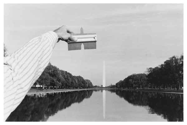 Black and white photo of the Washington Monument. From the bottom left hand corner a left hand holds up a tool matching the top of the monument.