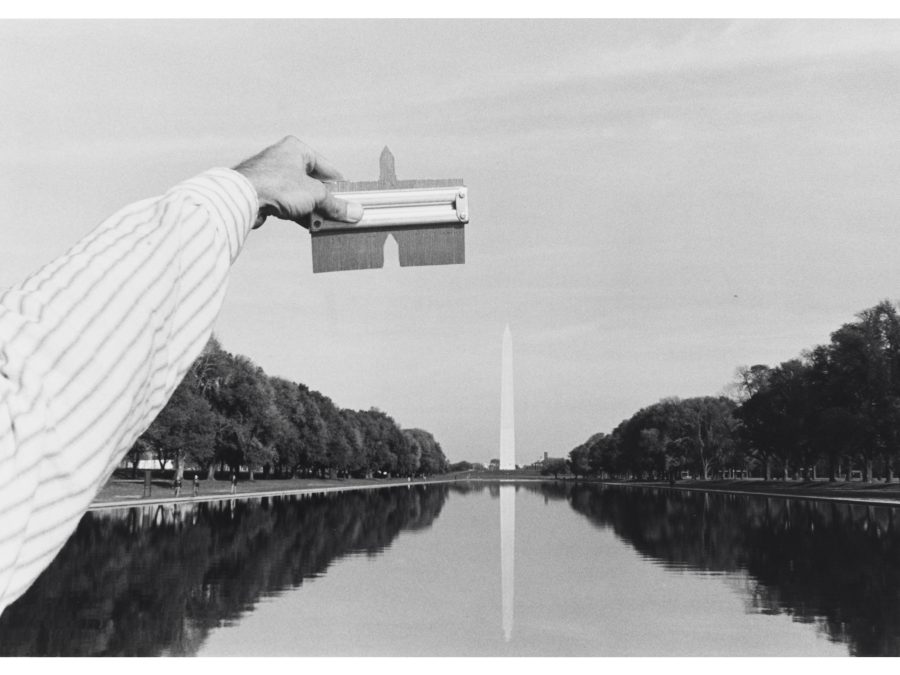 Black and white photo of the Washington Monument. From the bottom left hand corner a left hand holds up a tool matching the top of the monument.