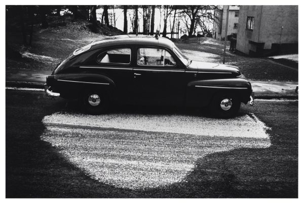 Black and white photo of a car's shadow that has blocked the sun causing the snow to melt everywhere else, creating the illusion of a white shadow on the road