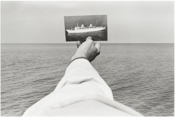 A black and white photo of a light skinned right arm wearing a white long-sleeved shirt holds a picture of a boat out over a body of water
