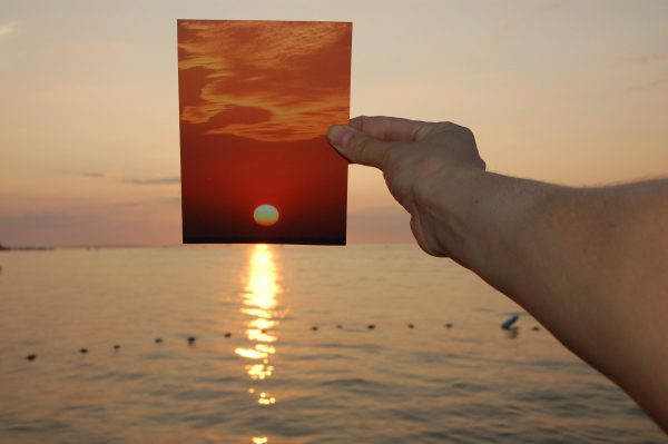 A light-skinned arm holds a picture of an orange sunset over a setting sun that is reflecting into a body of water