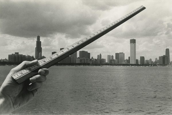 A light-skinned left hand holds a ruler diagonally across the Chicago skyline from the lake