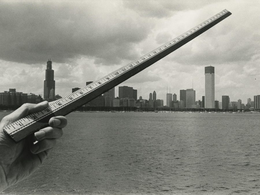 A light-skinned left hand holds a ruler diagonally across the Chicago skyline from the lake