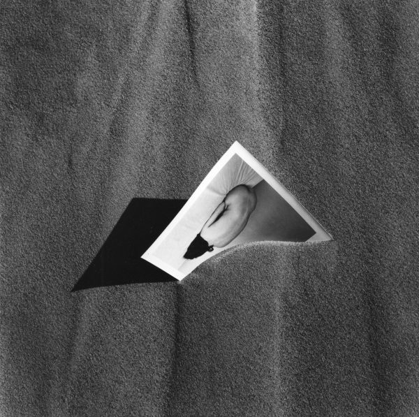 A black and white photograph of a picture of a nude person in a fetal position sticking out of a pile of sand
