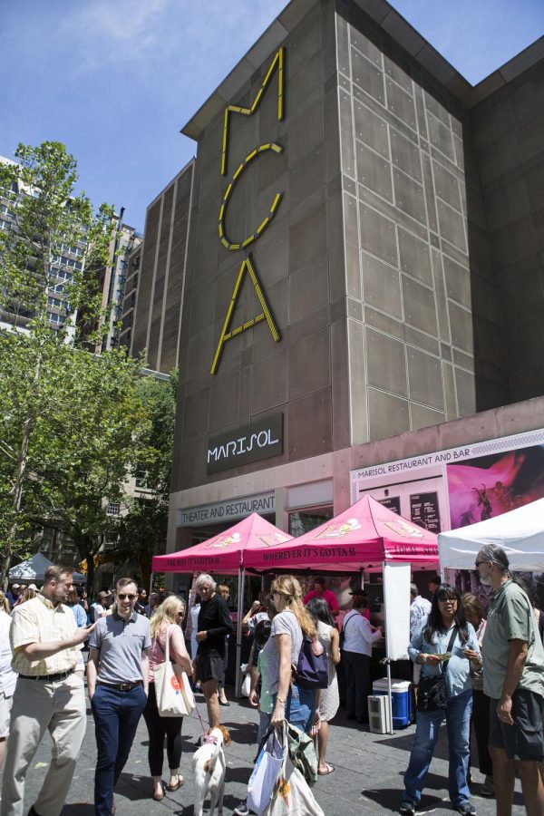 A large group of people walk past vendor tents in front of a large building with a light sign reading