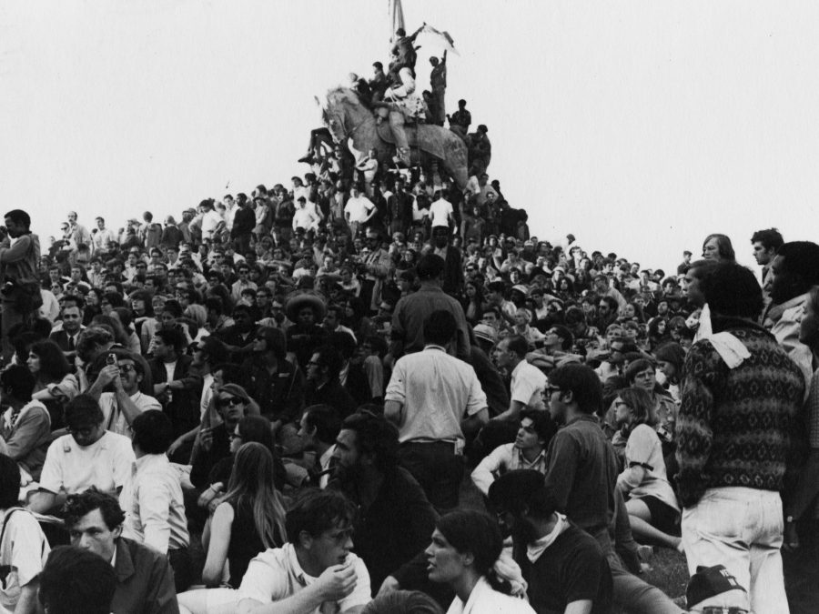 A black-and-white image shows a dense crowd of diverse young people gathered around and upon an elevated statue of a horse. Some people are climbing the horse, with the person at the top waving a flag.
