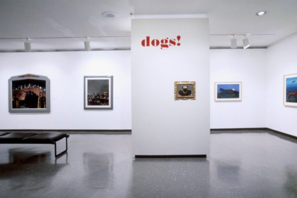 A modern art gallery room features various framed paintings on white walls. A central pillar displays the word 