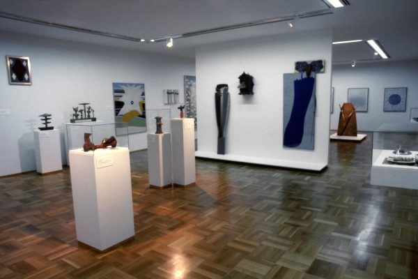 Gallery shot of sculptural artworks on pedestals and framed prints installed in white walled gallery with wooden parquet floors.