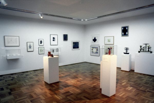 Gallery shot of sculptural artworks on pedestals and framed prints installed in white walled gallery with wooden parquet floors.