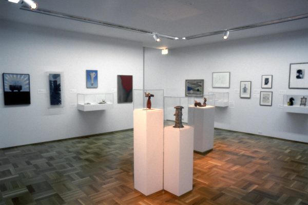 Gallery shot of sculptural artworks on pedestals and framed prints installed in white walled gallery with wooden parquet floors.