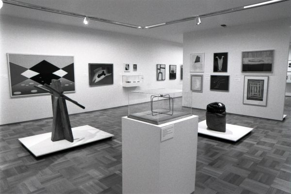 Gallery shot of sculptural artworks on pedestals and framed prints installed in white walled gallery with wooden parquet floors.