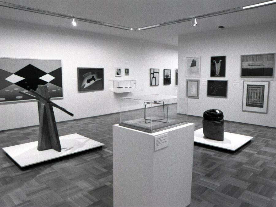 Gallery shot of sculptural artworks on pedestals and framed prints installed in white walled gallery with wooden parquet floors.