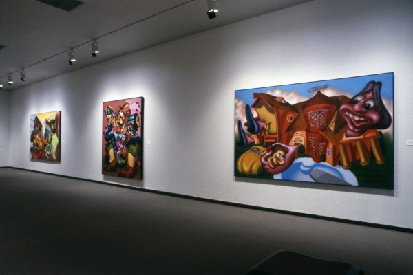 Three large, colorful paintings hang on a dramatically lit white wall.