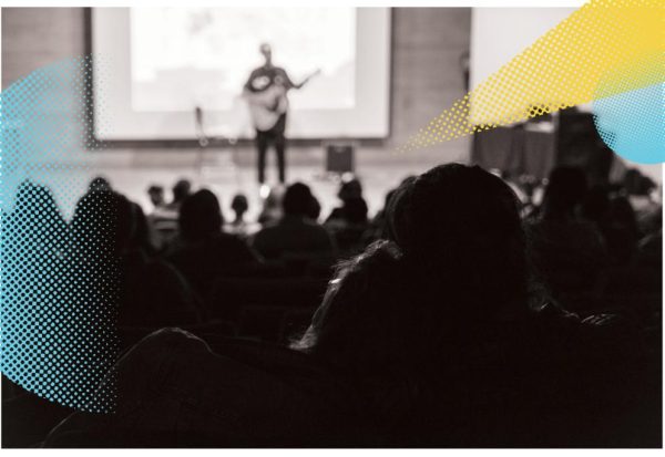 A black and white photograph of two audience members watching a man play a guitar on a stage is overlaid with geometric blue and yellow designs.