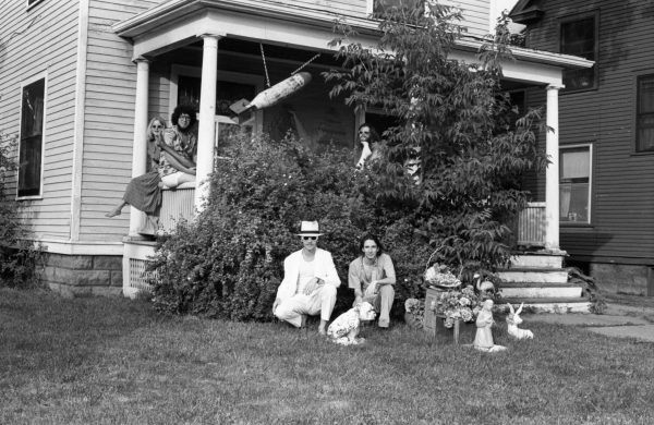 Black-and-white photo of a group of people and a dog pose in front of a house with overgrown plants