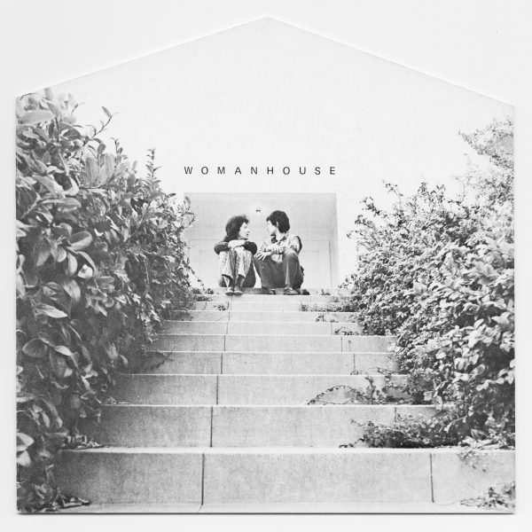 Two people sit at the top of an outdoor set of stairs above them is a white building with the entrance labeled Womanhouse