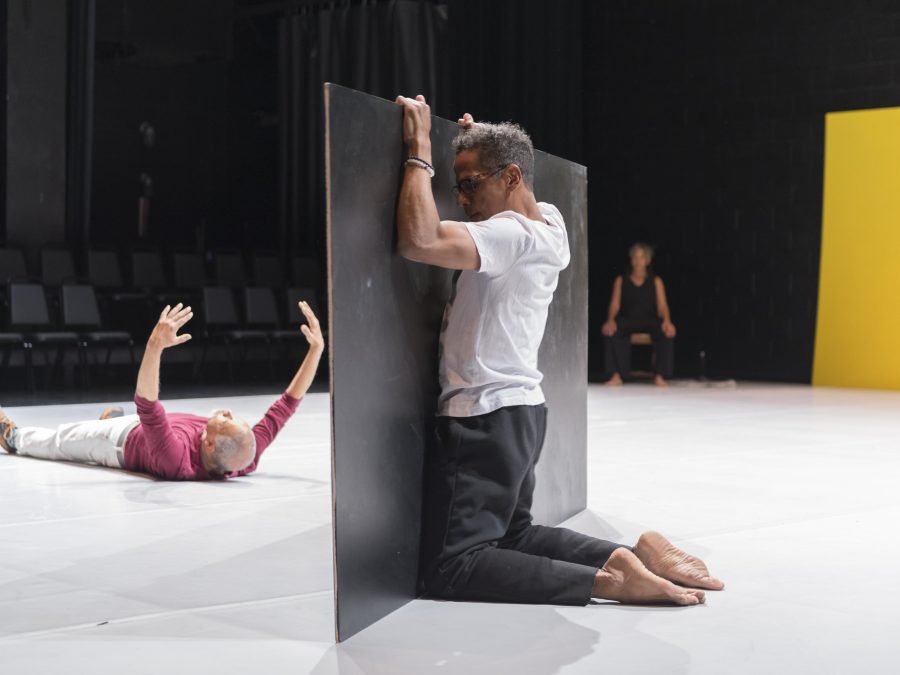 Three people appear on a stage: one hides behind a board, behind which another rolls on the round, hands up in the air, as the third looks on from the wings