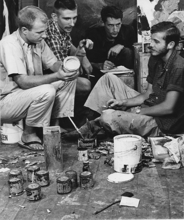 Black and white photo of four men sitting on the ground and on paint pails surrounded by art-making supplies