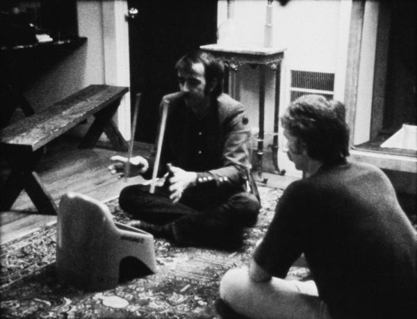 Archival image of two people sitting on a rug near an object that looks like a plastic stool. One of them holds a pair of drumsticks.